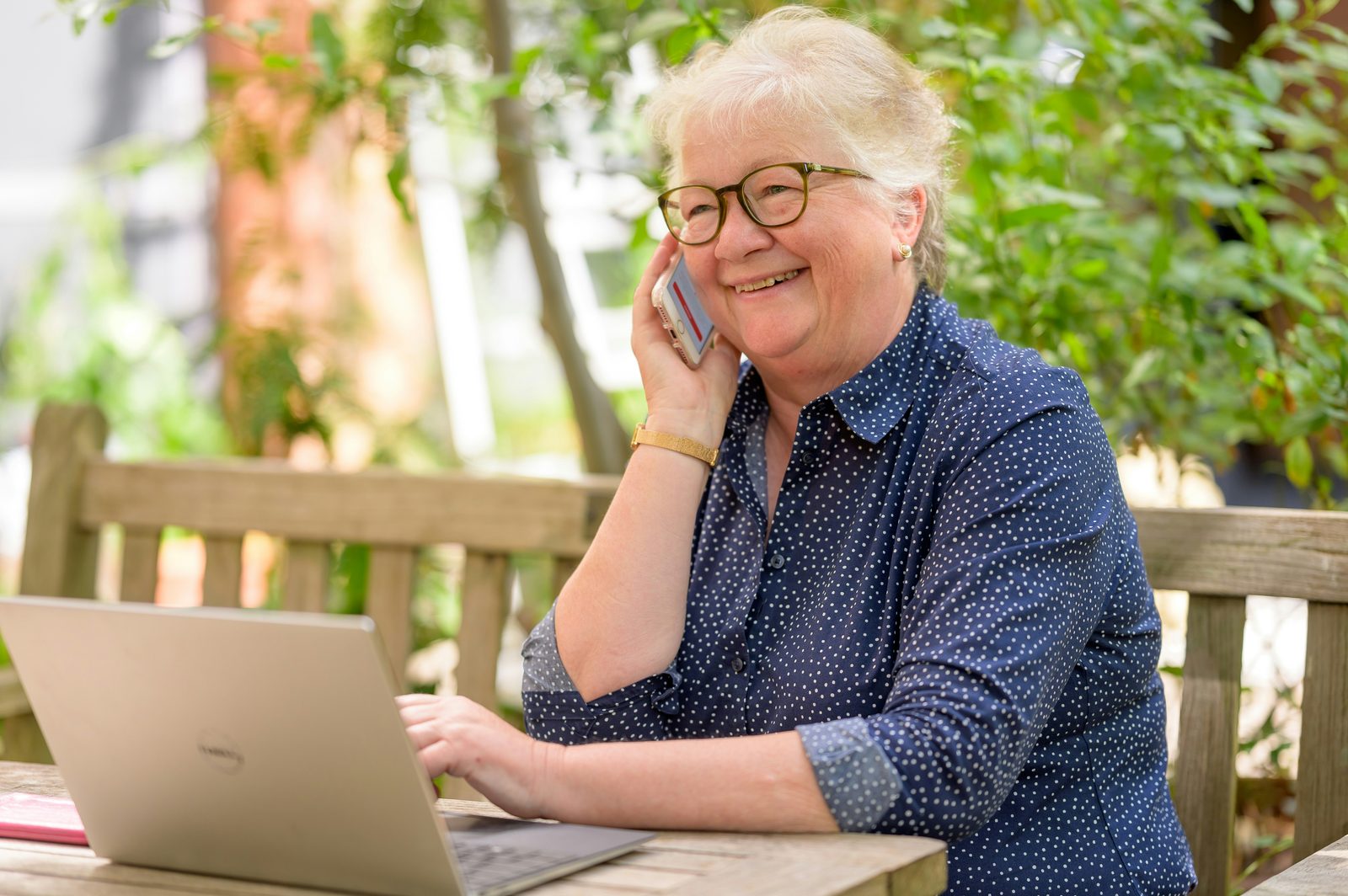 Smiling senior woman on a phone call outdoors with her laptop